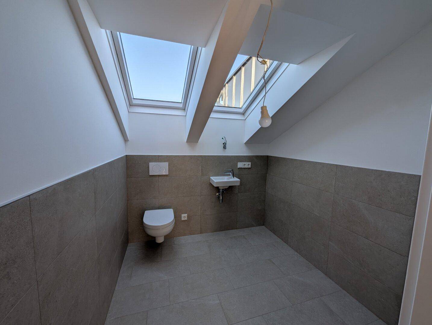 Kleines WC mit Waschbecken, Toilette, grauen Fliesen und Dachfenstern für natürliches Licht.