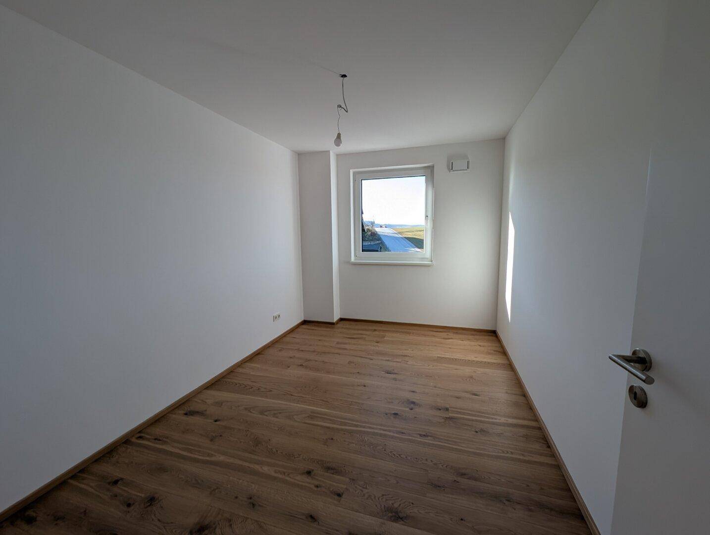 Helles, leeres Zimmer mit Holzboden und Fenster, geeignet als Kinder- oder Gästezimmer.