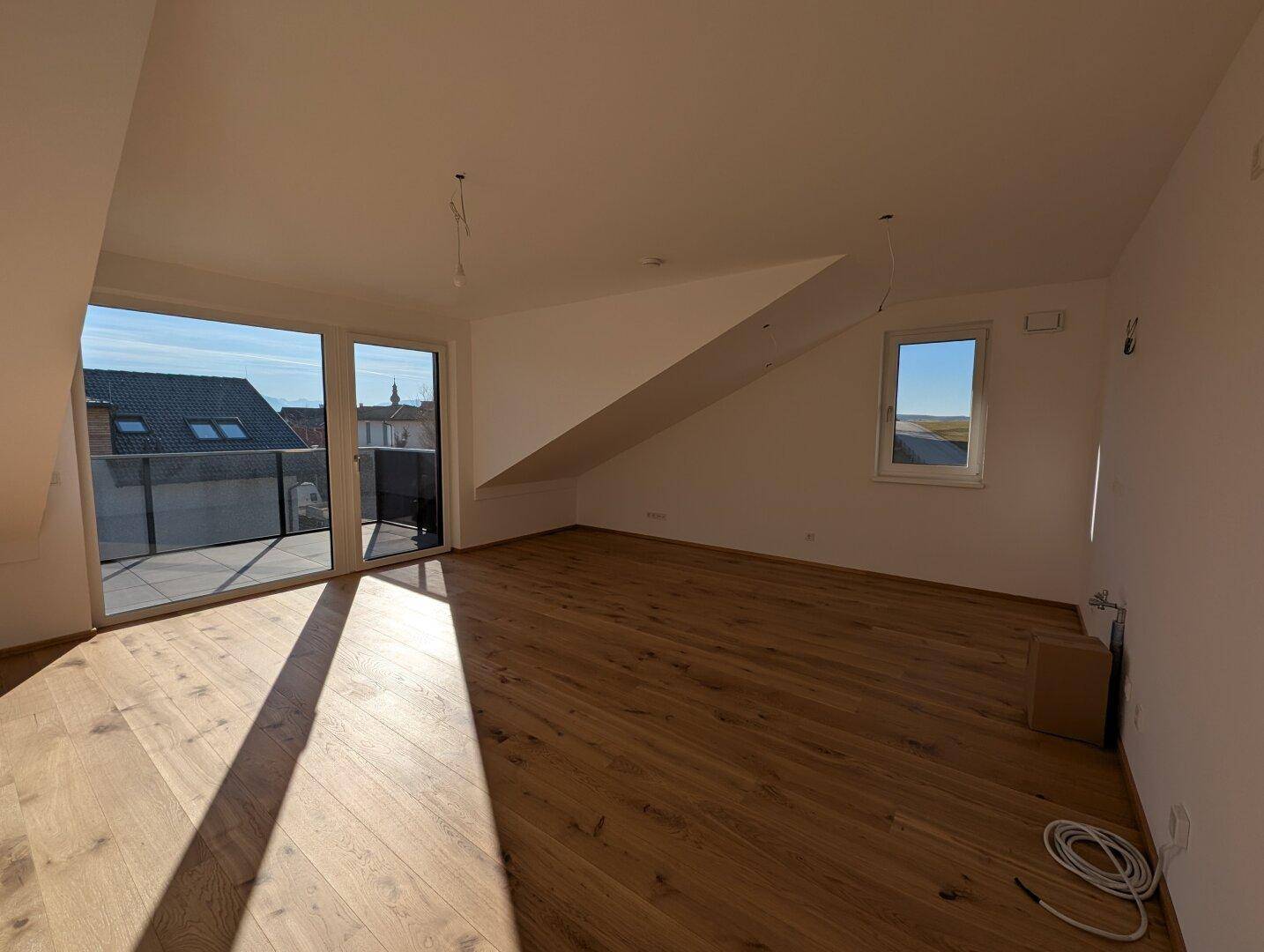 Helles, leeres Wohnzimmer mit Holzboden, großen Schiebetüren zum Balkon und Dachschrägen.