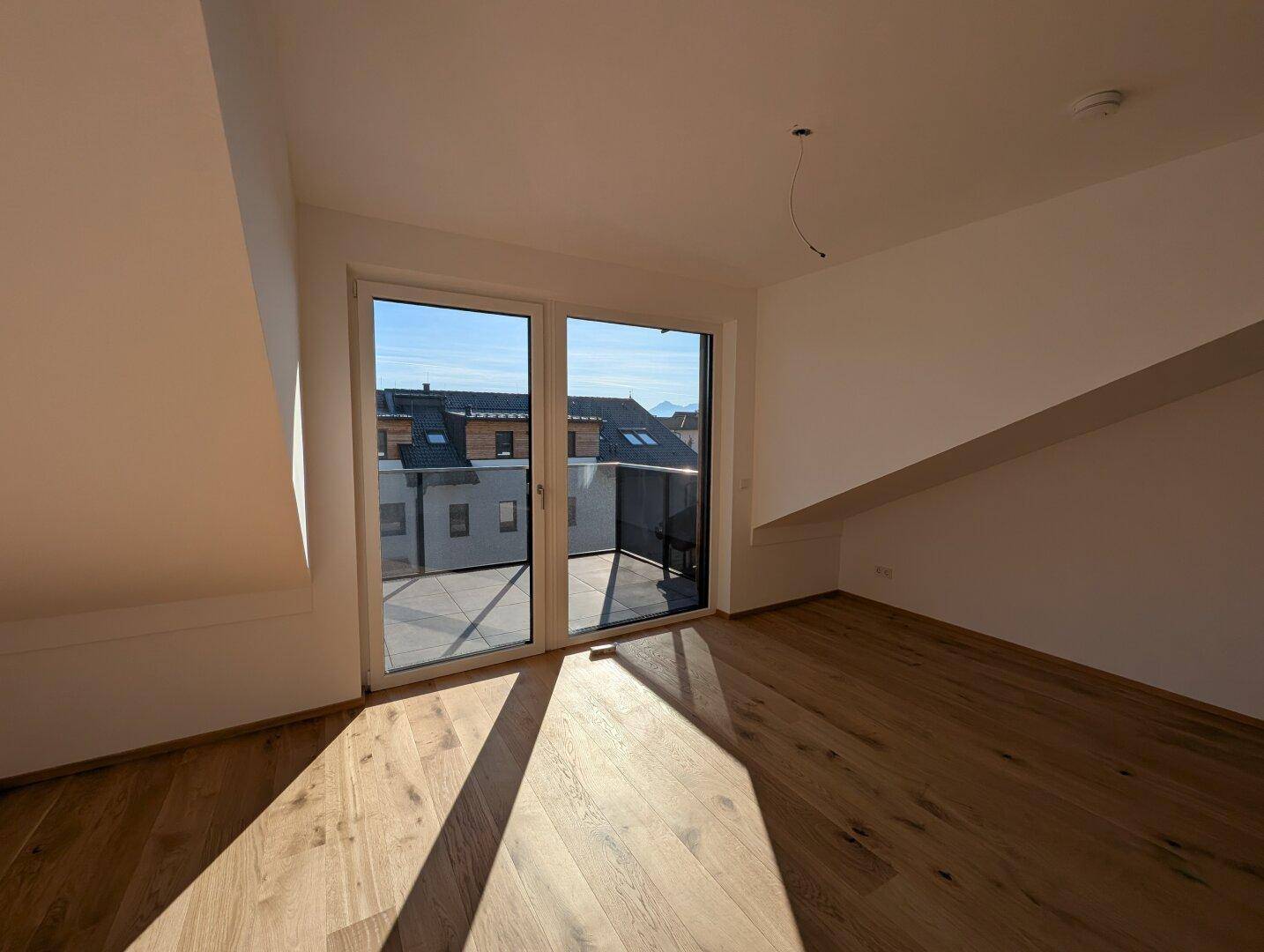 Lichtdurchfluteter Raum mit Holzboden und großen Fenstern, die auf einen Balkon führen.