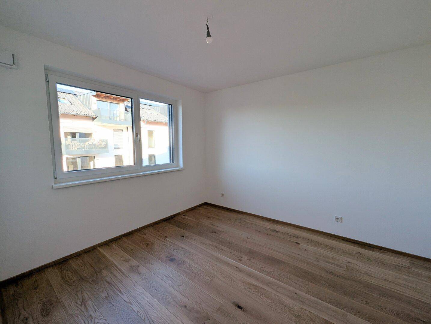 Ruhiges Schlafzimmer mit hellem Parkettboden und einem großen Fenster, das viel Tageslicht hereinlässt.