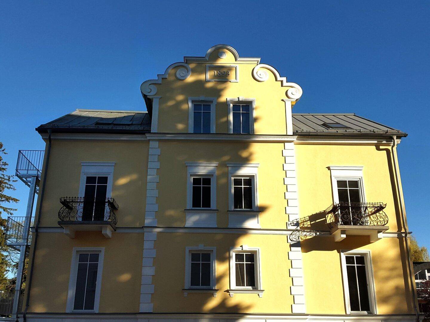 Vorderansicht des historischen Gebäudes mit gelber Fassade, dekorativen Balkonen und Satteldach.