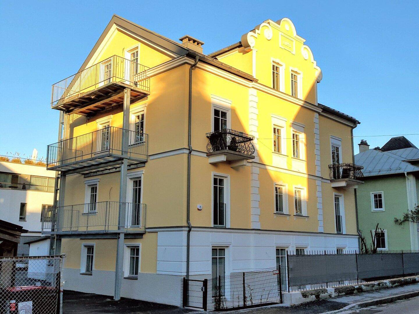Rückansicht des Gebäudes mit gelber Fassade und einem modernen Anbau mit mehreren Balkonen.