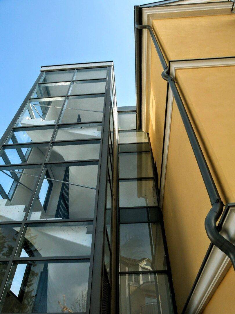 Moderner Glasaufzug neben der historischen gelben Fassade des Gebäudes, unter blauem Himmel.