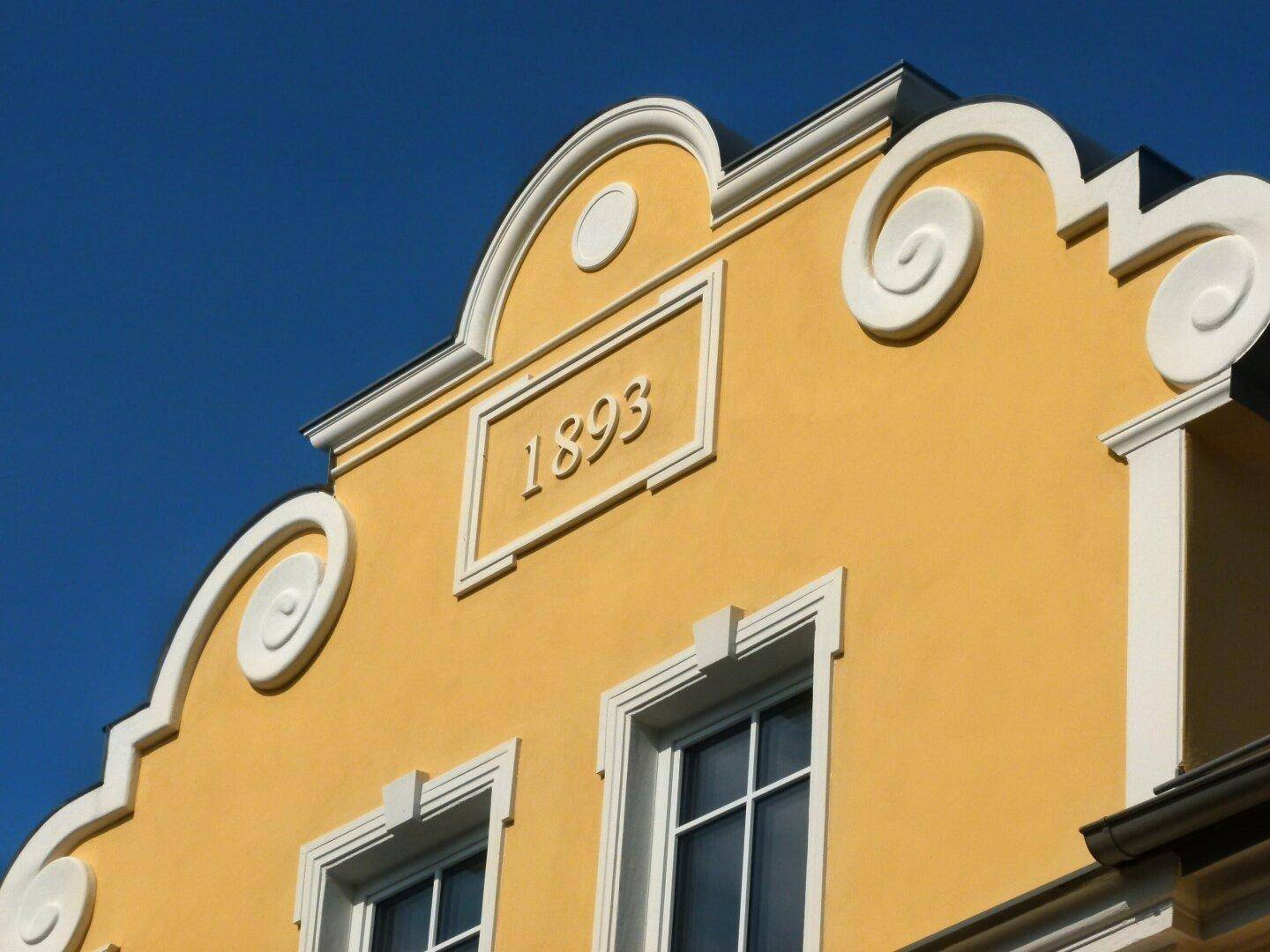 Detailaufnahme des verzierten Giebels mit der Jahreszahl 1893 und weißem Stuck an der gelben Fassade.