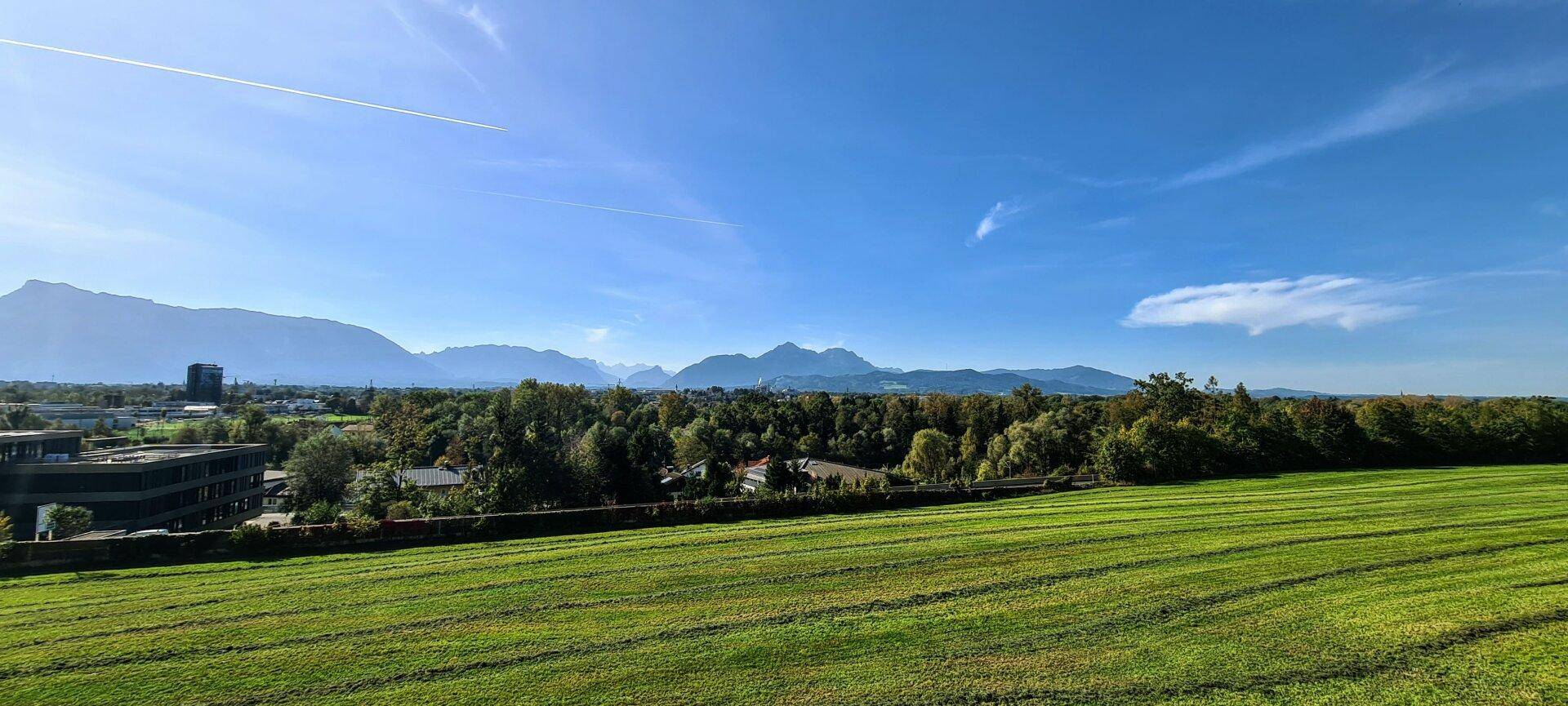 Weitläufiger Panoramablick über grüne Wiesen, Gebäude und eine beeindruckende Bergkulisse.