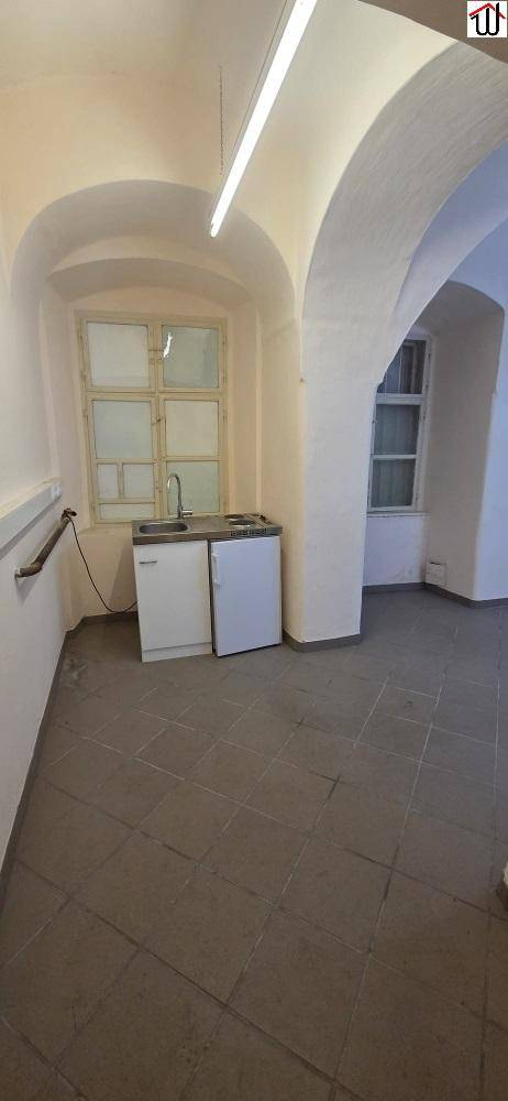 Heller Raum mit Gewölbedecken, grauen Fliesen und einer kleinen Küchenzeile mit Spüle und Kühlschrank.