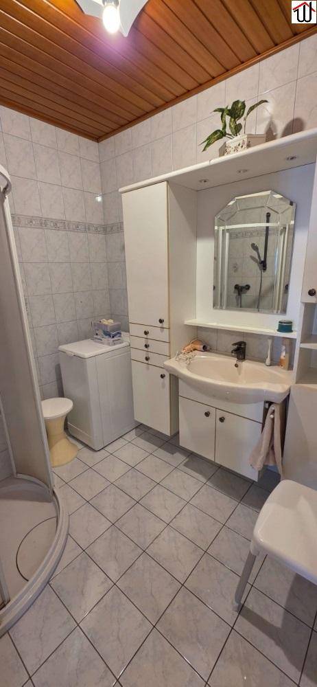 Funktionales Badezimmer mit Dusche, Waschbecken und Waschmaschine, gefliesten Wänden und Holzdecke.