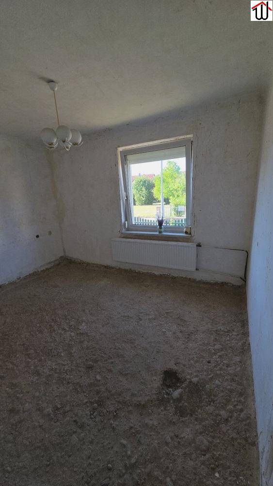 Leerer Raum in Renovierung mit unfertigem Boden, weißen Wänden und einem Fenster mit Blick ins Grüne.
