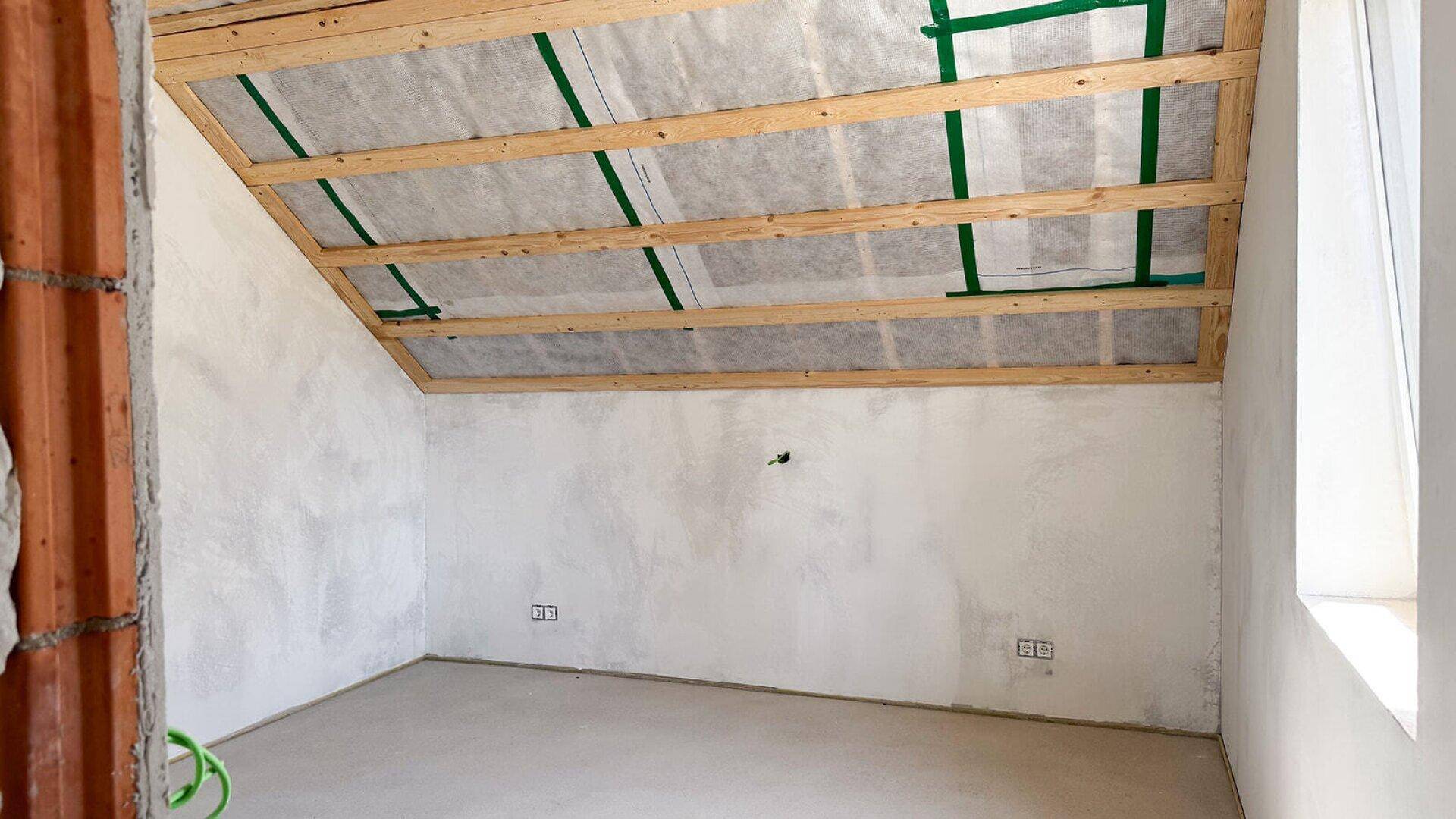 Rohbau eines Dachzimmers mit sichtbaren Holzbalken und einem Fenster, wartet auf Fertigstellung.