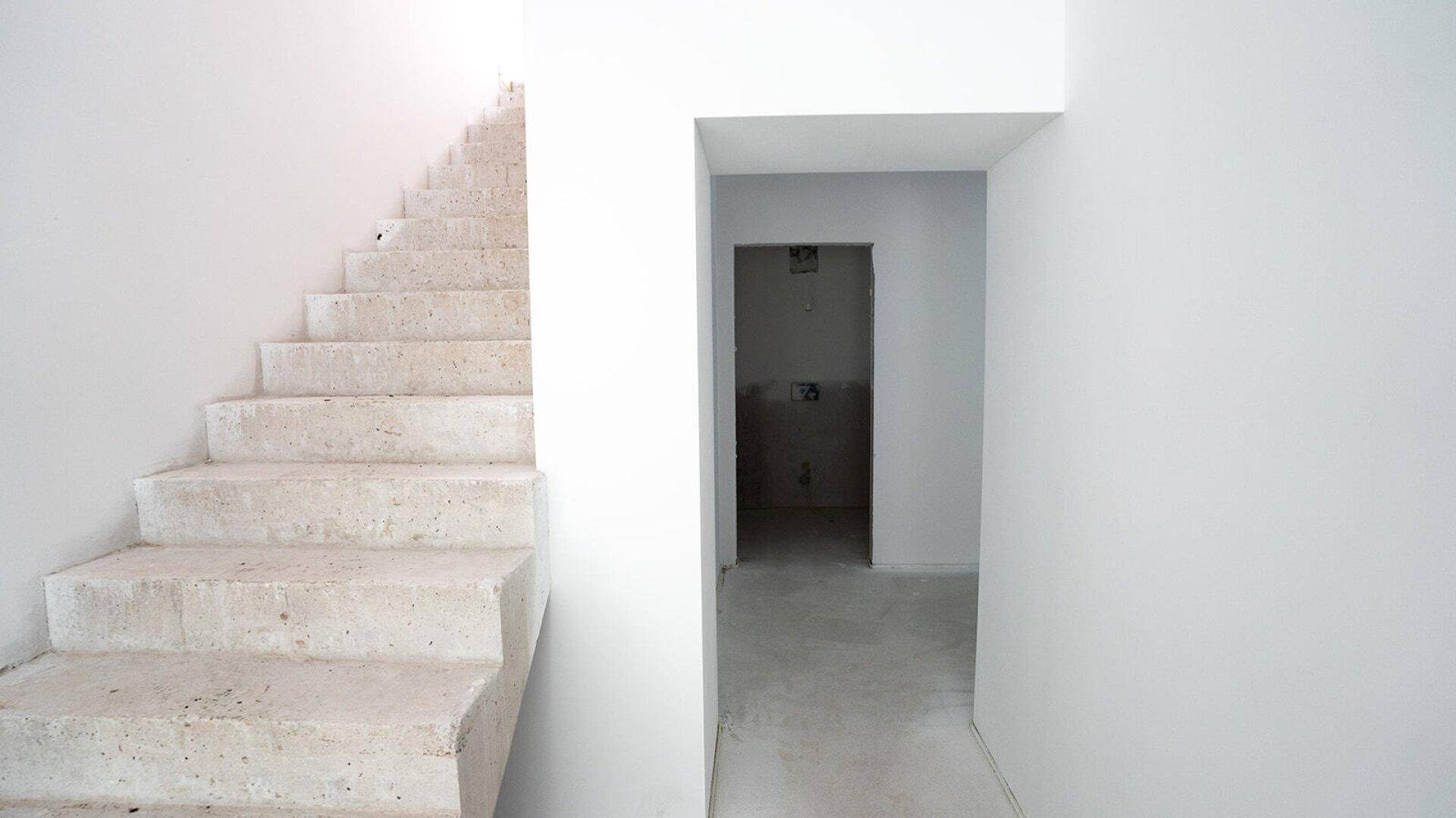 Treppenaufgang im Rohbau neben einem Türrahmen, der zu einem weiteren Raum führt.
