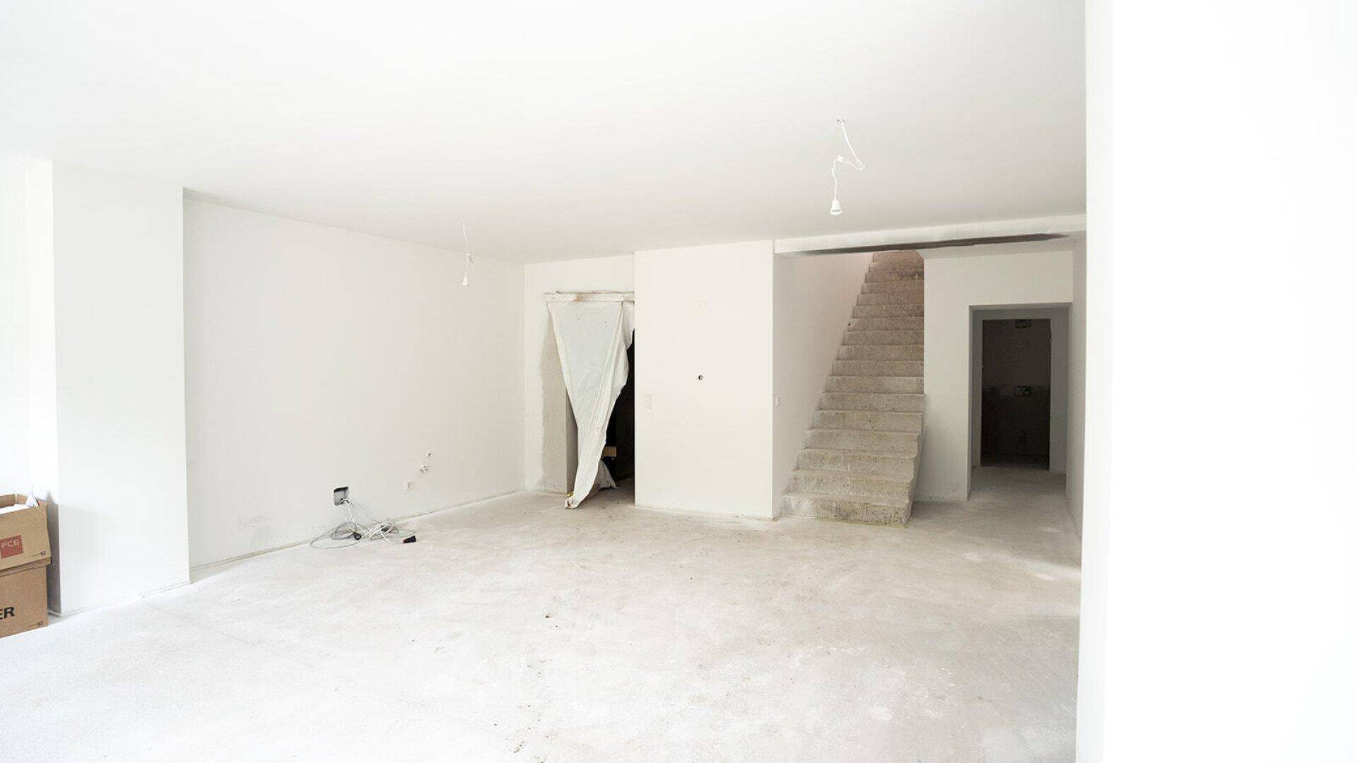 Großer, heller Raum im Rohbau mit sichtbarem Treppenaufgang und weißen Wänden.