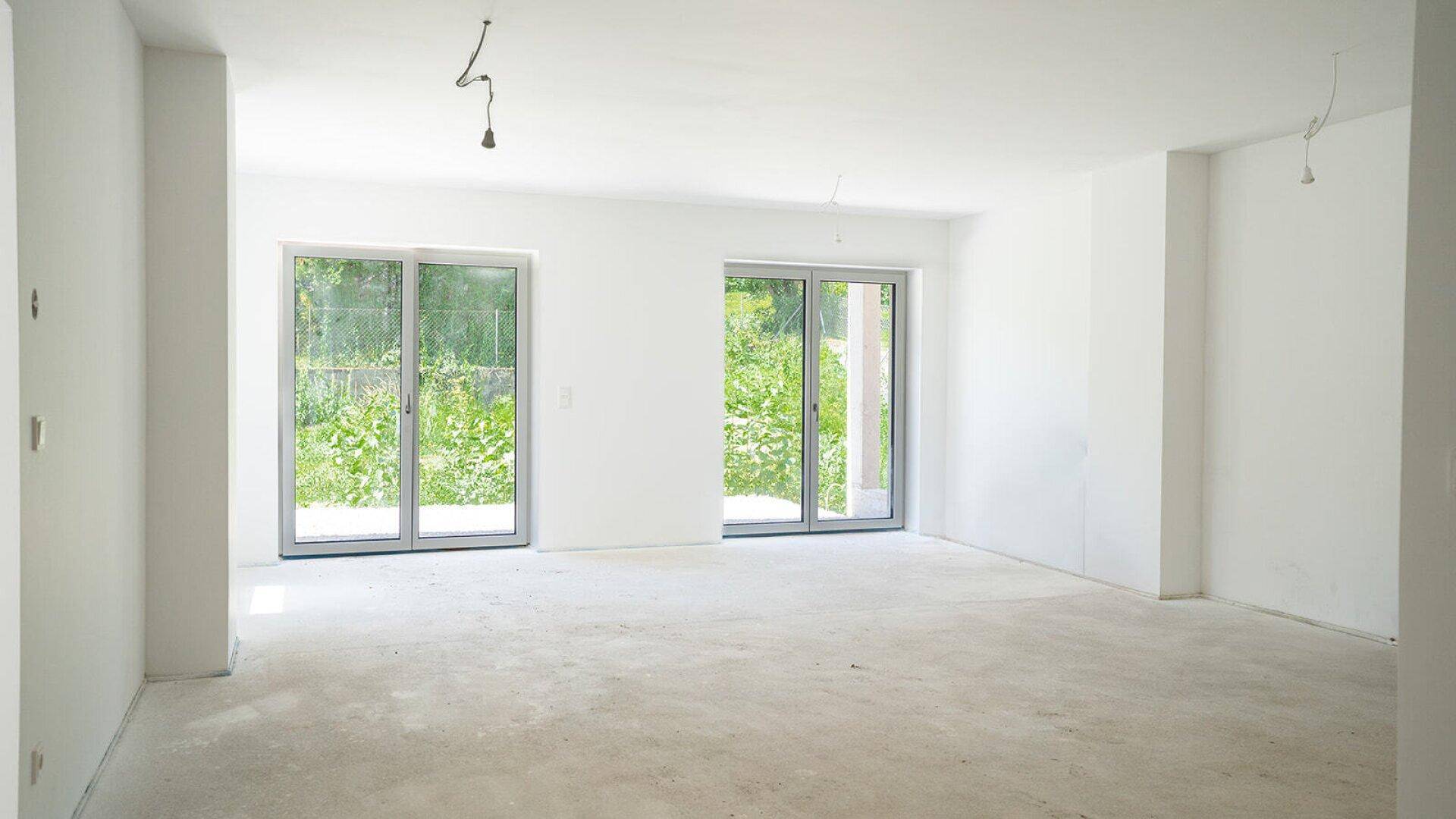 Heller, unmöblierter Raum mit großen Fenstertüren und Blick ins Grüne.