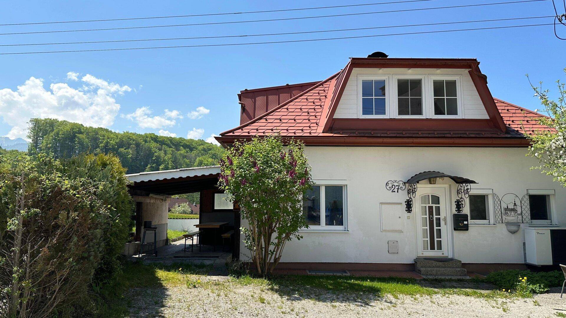 Freundliche Vorderansicht des Hauses mit rotem Dach, weißer Fassade und einem Carport bei blauem Himmel.