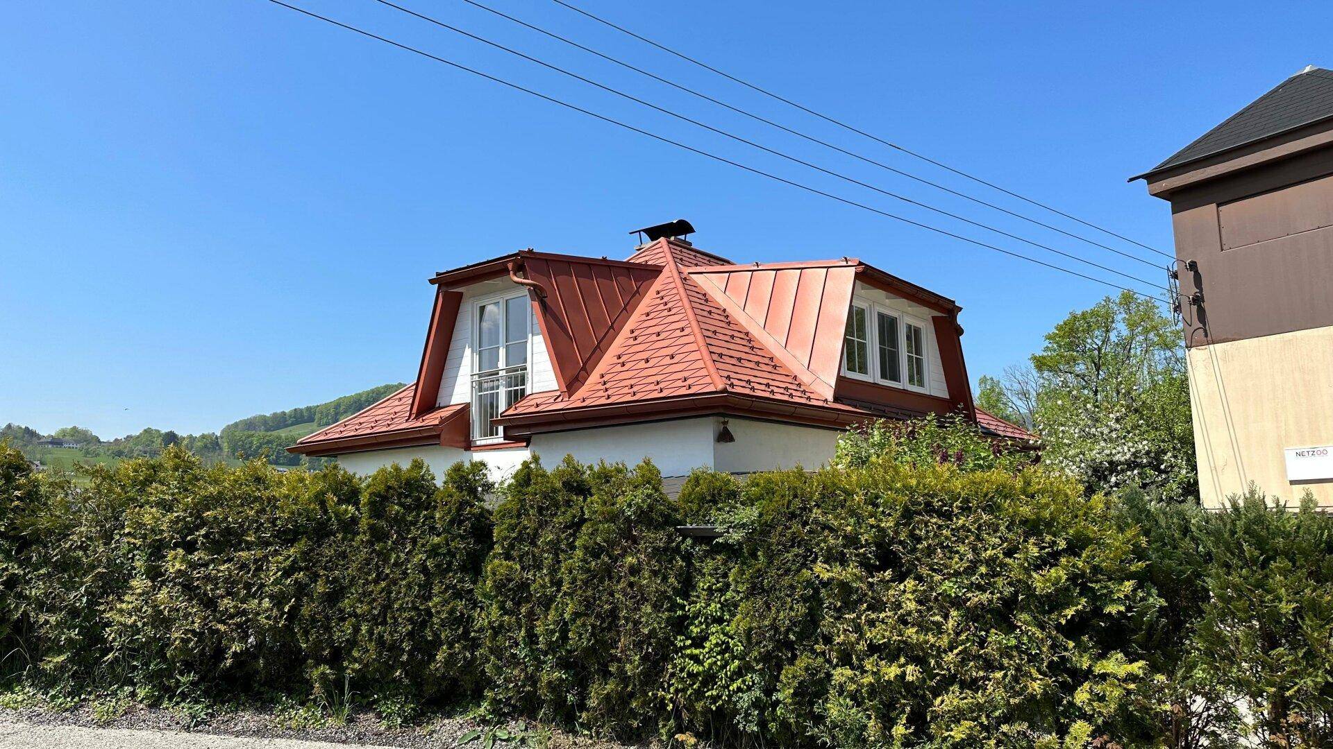 Seitenansicht des Hauses, teilweise von einer hohen Hecke verdeckt, unter blauem Himmel.