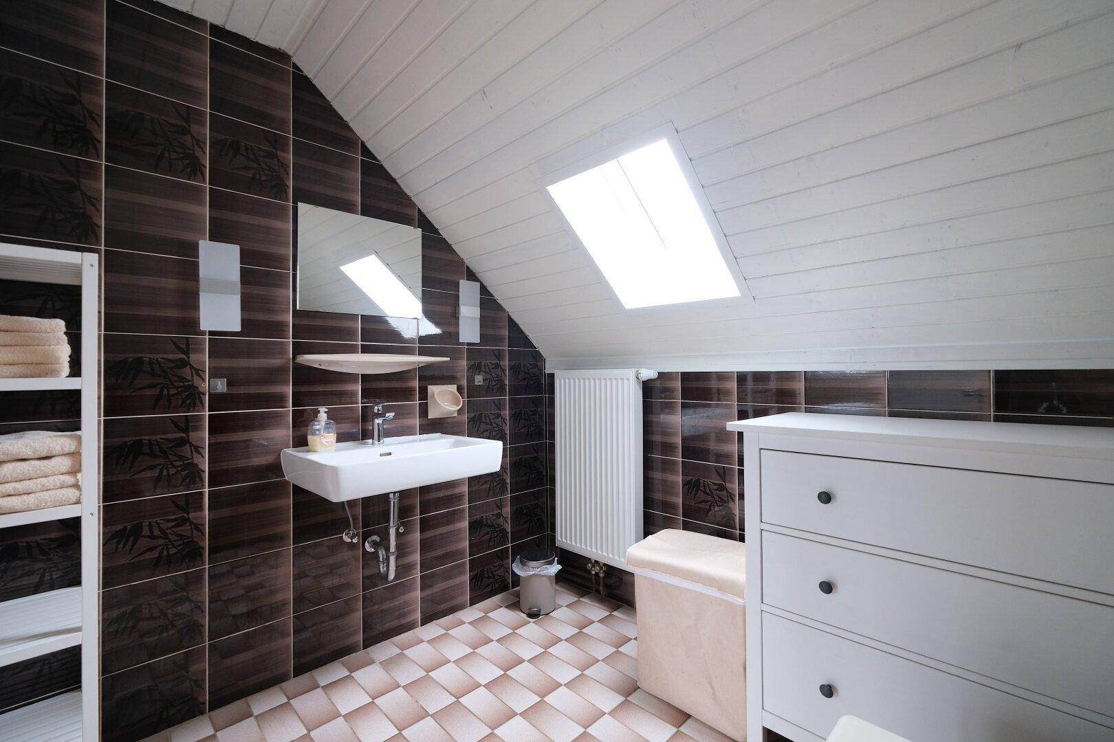 Badezimmer im Dachgeschoss mit dunklen Wandfliesen, hellem Boden und einem Dachfenster für natürliches Licht.
