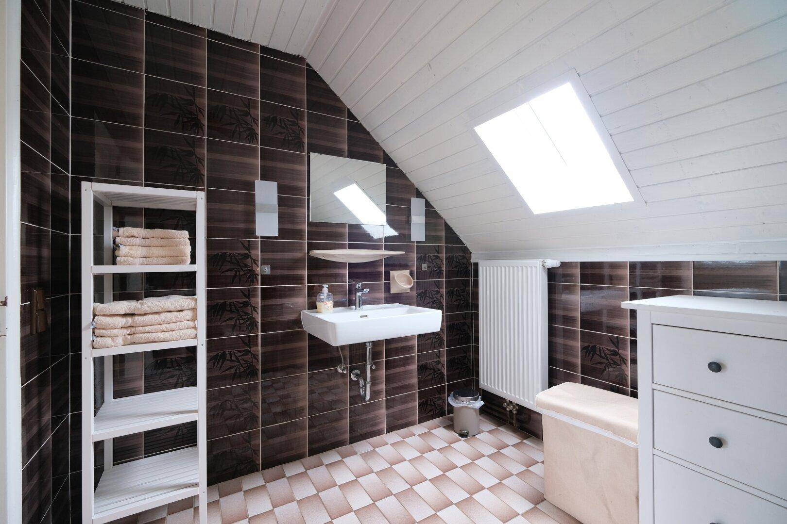 Helles Badezimmer im Dachgeschoss mit braunen Wandfliesen, kariertem Boden und praktischen Regalen.