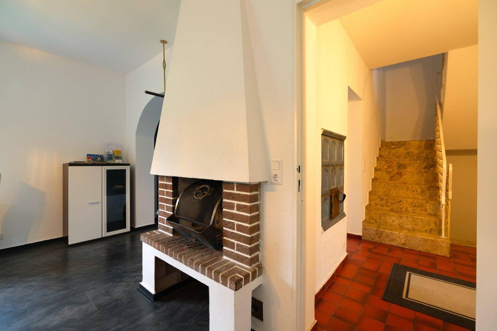 Wohnzimmer mit rustikalem Ziegelkamin und dunklem Boden, angrenzend an einen Flur mit Treppe.