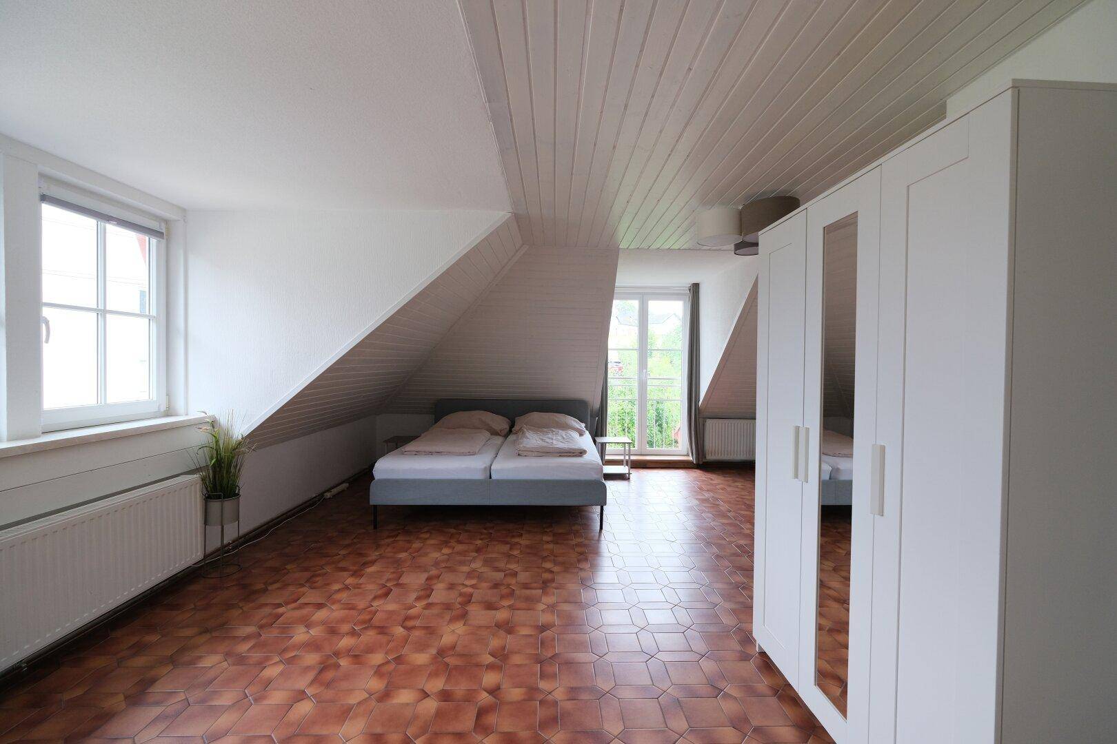 Dachgeschoss-Schlafzimmer mit Fenster, Heizkörper und schrägen Wänden, bietet viel Tageslicht.