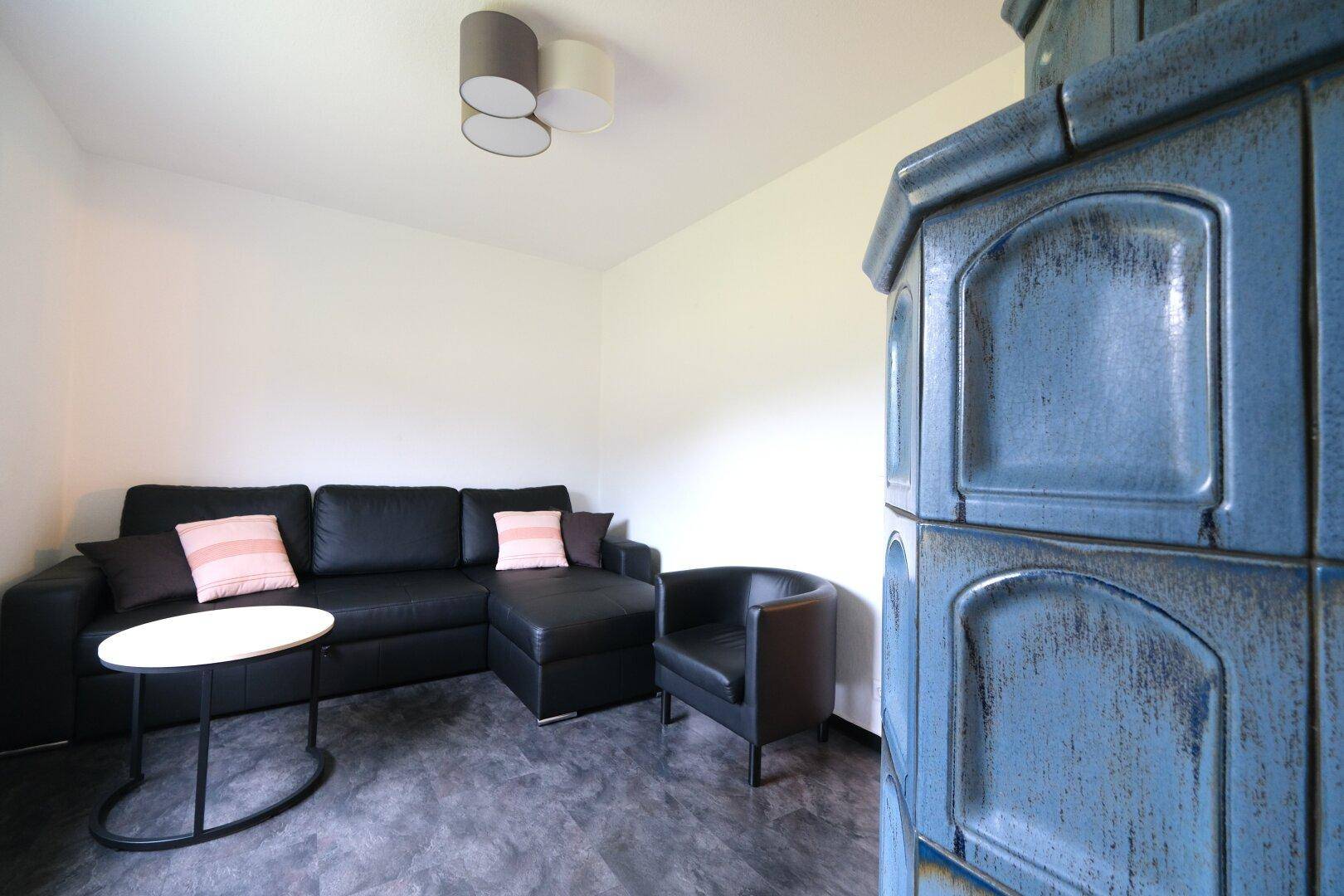 Gemütliches Wohnzimmer mit einem blauen Kachelofen, schwarzem Sofa und einem bequemen Sessel.