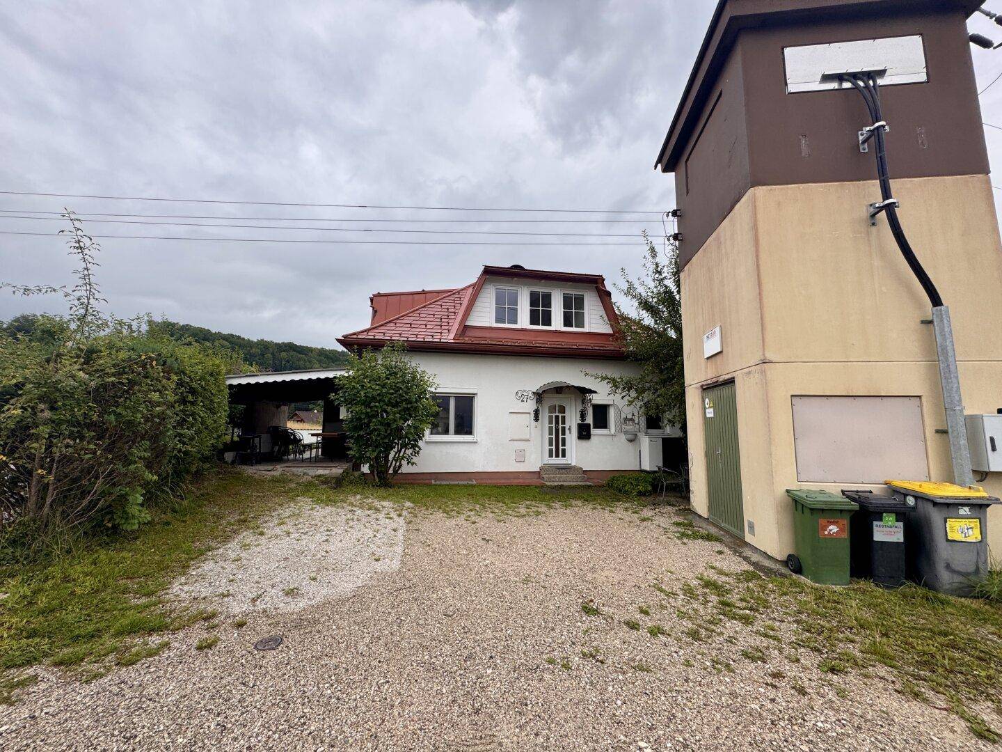 Vorderansicht des Hauses mit weißer Fassade, rotem Dach und Kiesauffahrt unter bewölktem Himmel.