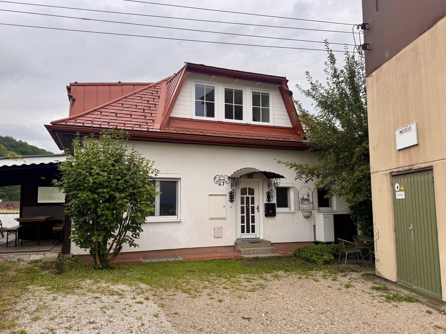 Vorderansicht des Hauses mit rotem Dach, weißer Fassade und Kiesauffahrt unter bewölktem Himmel.