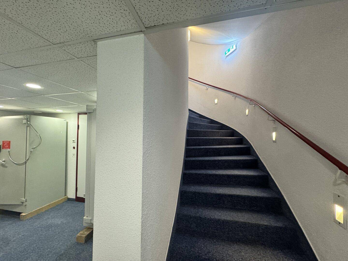 Geschwungene Treppe mit Teppichboden und Handlauf, die zu einem oberen Stockwerk führt, beleuchtet von Wandleuchten.