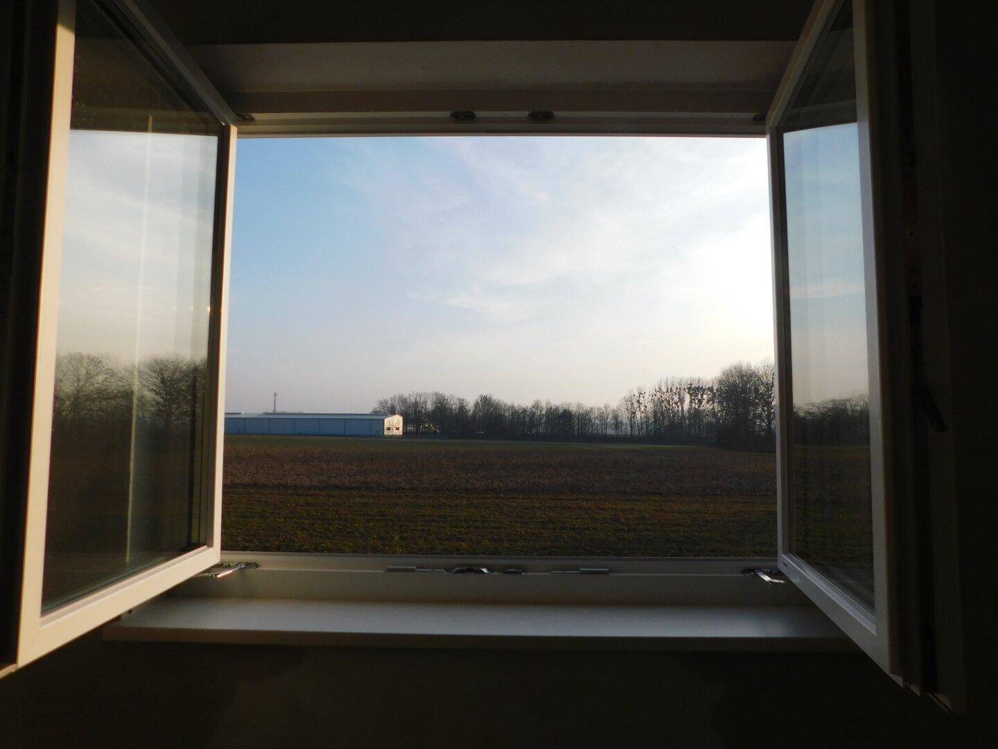Blick aus einem geöffneten Fenster auf eine weite Landschaft mit Feldern und Bäumen.