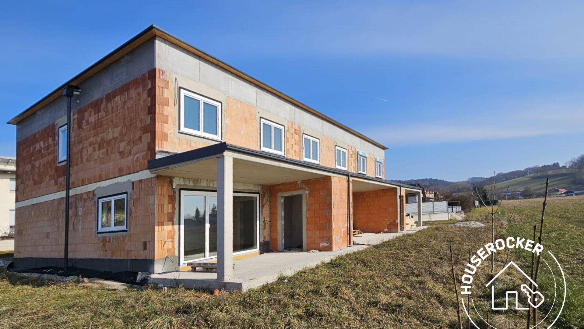 Rohbau eines Reihenhauses mit großen Fensteröffnungen und zukünftigen Terrassenbereichen in ländlicher Umgebung.