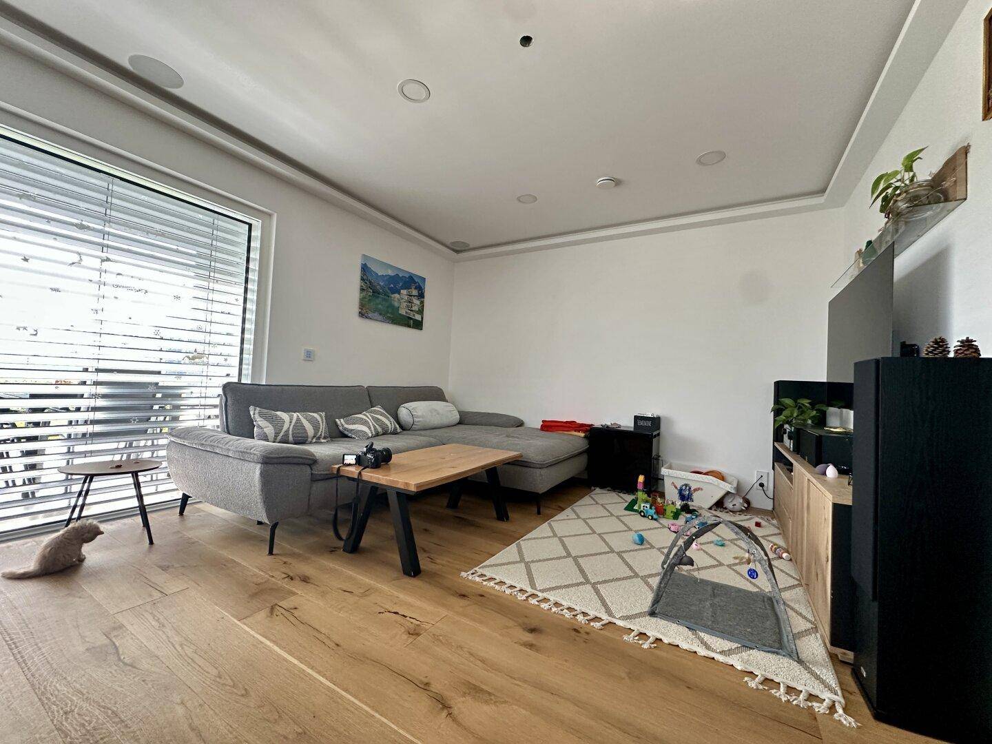 Gemütliches Wohnzimmer mit großem grauen Sofa, Holzboden und bodentiefen Fenstern.