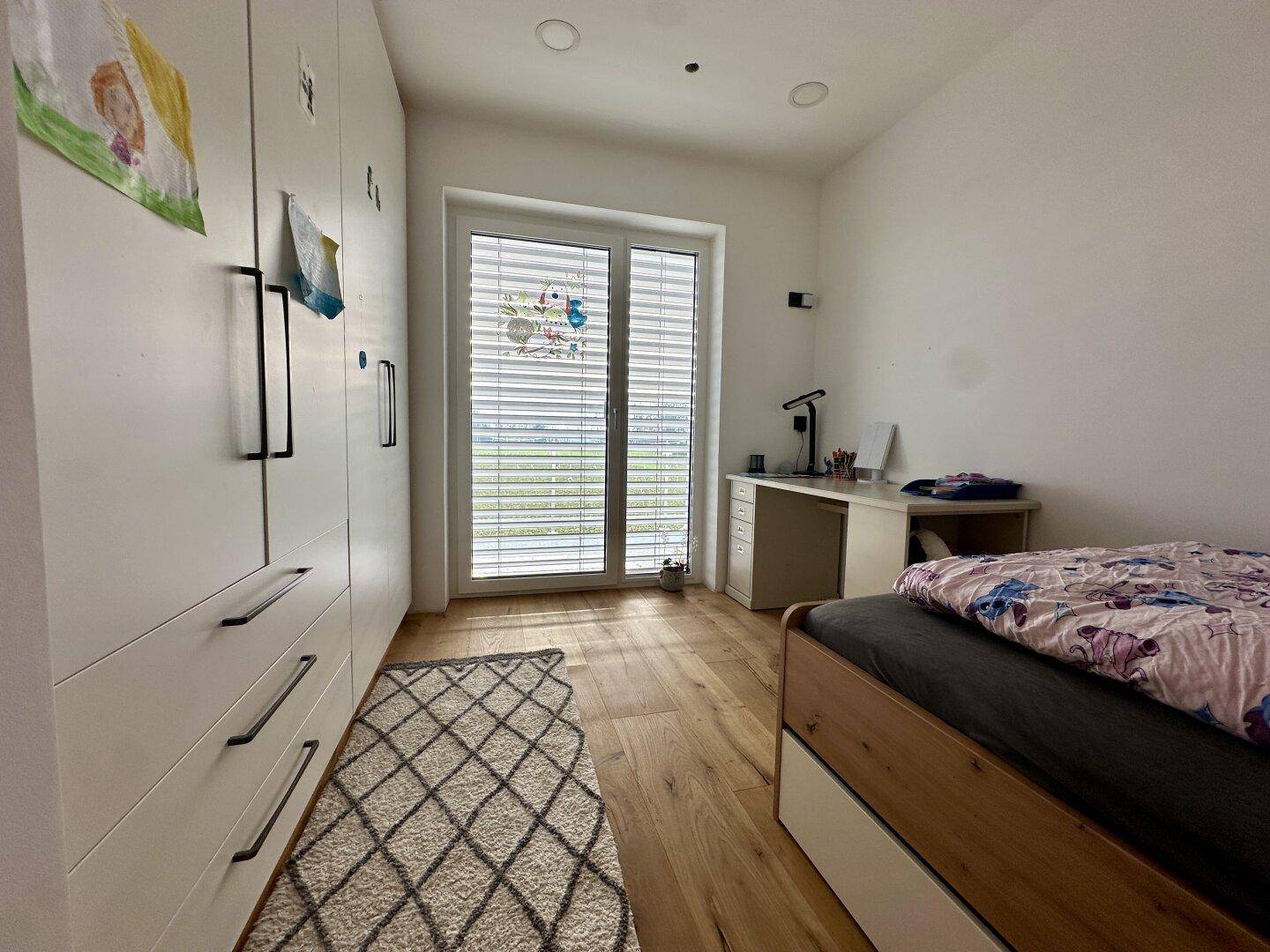 Kinderzimmer mit Bett, Schreibtisch, Einbauschrank und großem Fenster mit Blick ins Grüne.