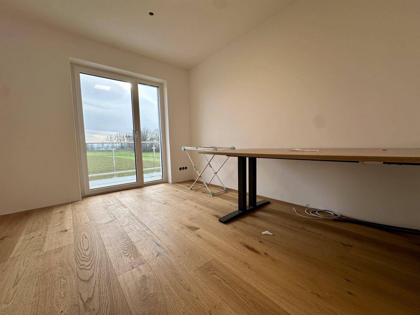 Heller Raum mit Holzboden und Zugang zu einem Balkon oder einer Terrasse mit Blick ins Grüne.