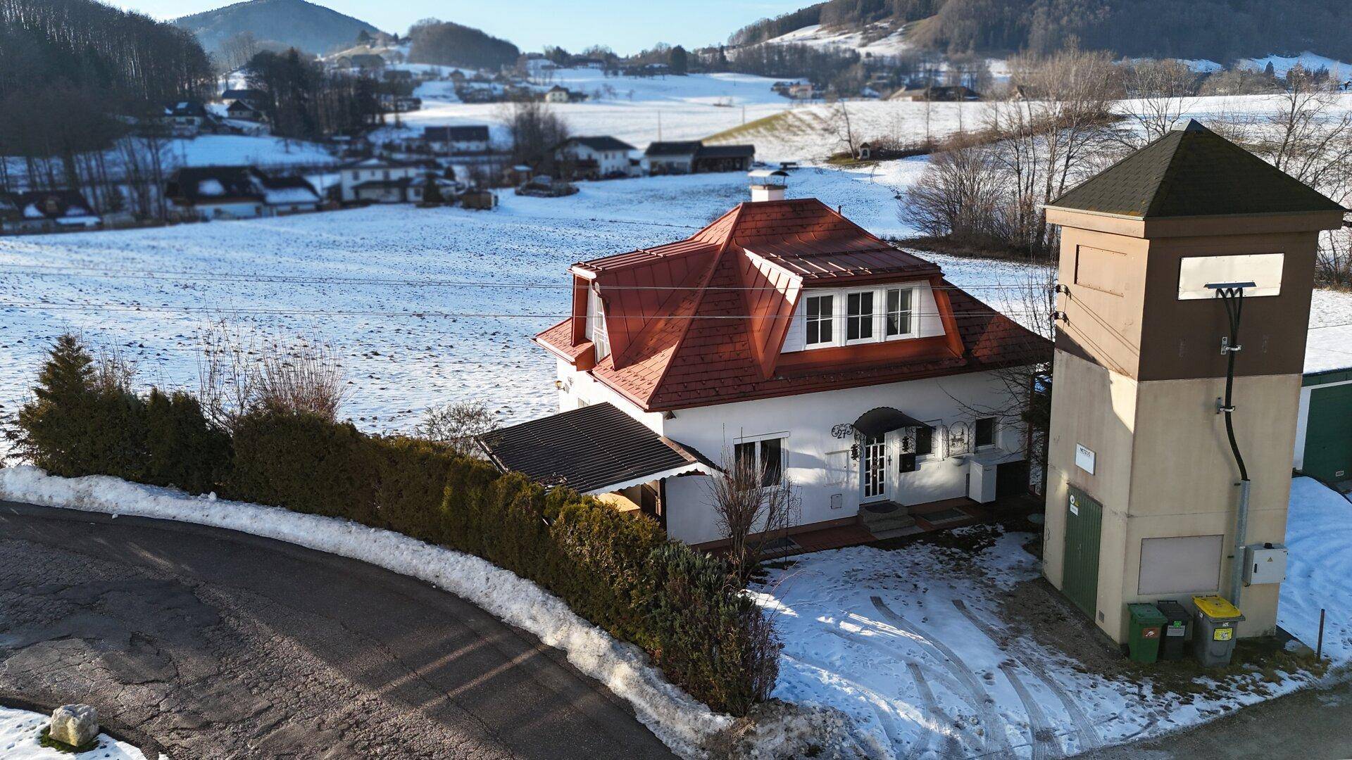 Luftaufnahme eines Hauses mit rotem Dach und verschneiter Umgebung im Winter.