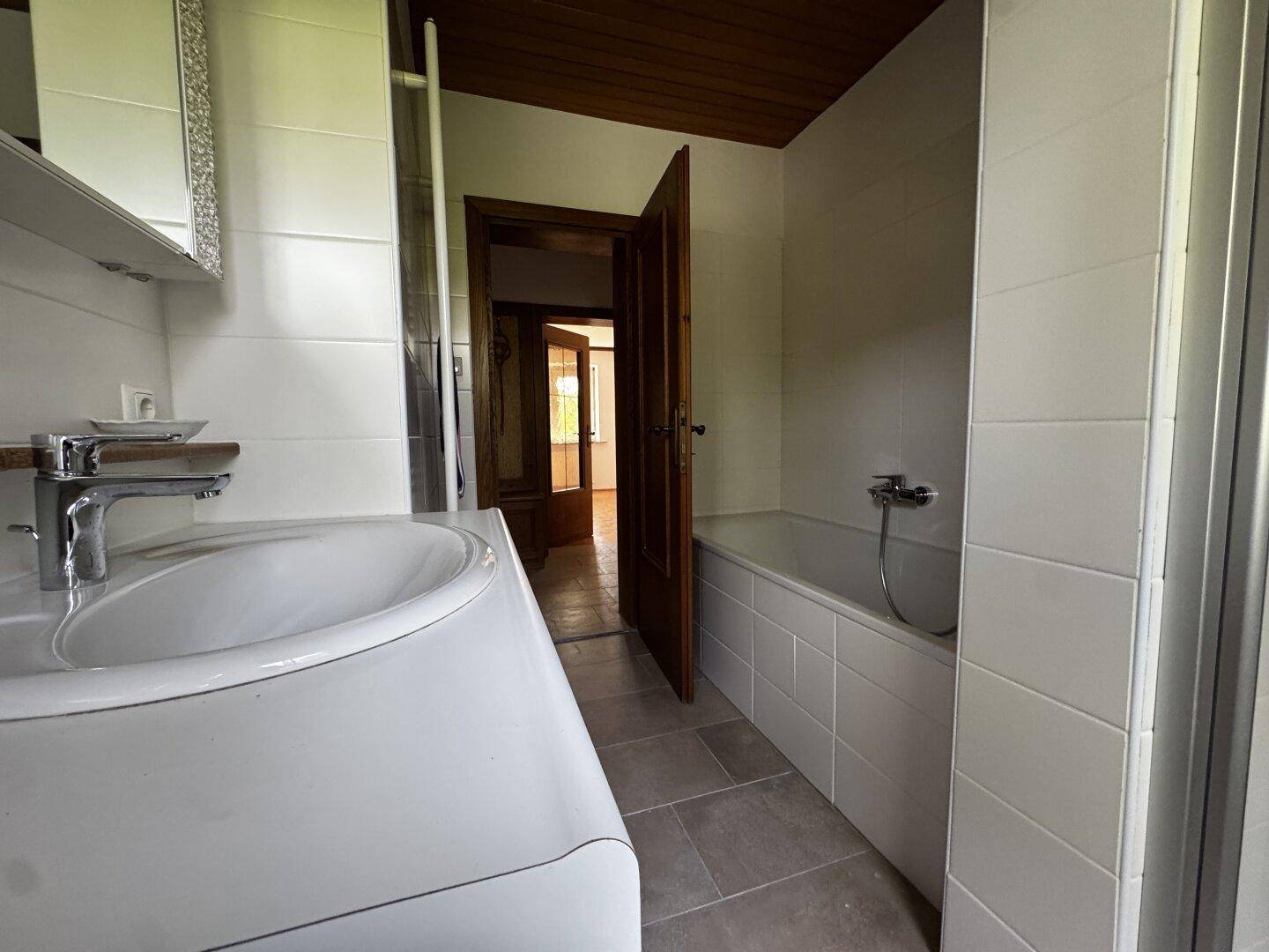 Helles Badezimmer mit Badewanne, Waschbecken und weißen Fliesen, Holzbalkendecke.