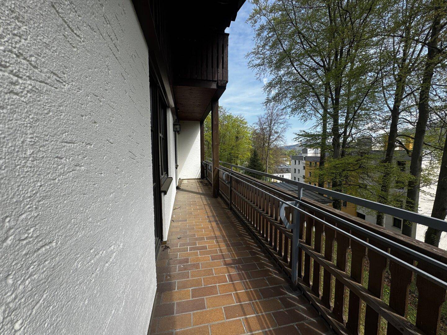 Langer Balkon mit Fliesenboden und Holzgeländer, umgeben von Bäumen.