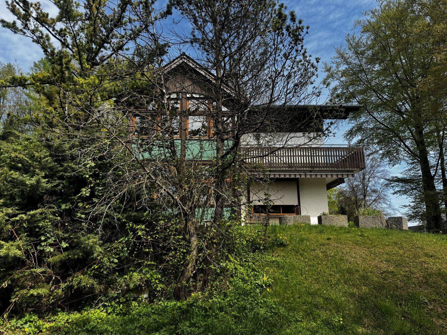 Hausfassade teilweise von Bäumen verdeckt, eingebettet in eine grüne Umgebung.