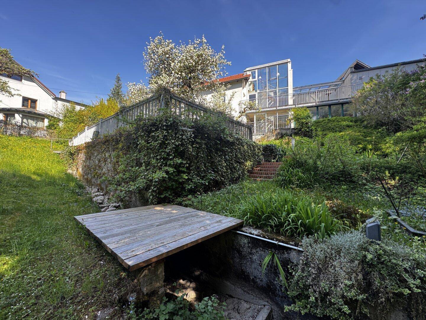 Naturbelassener Gartenbereich mit Holzbrücke und Blick auf das moderne Gebäude.