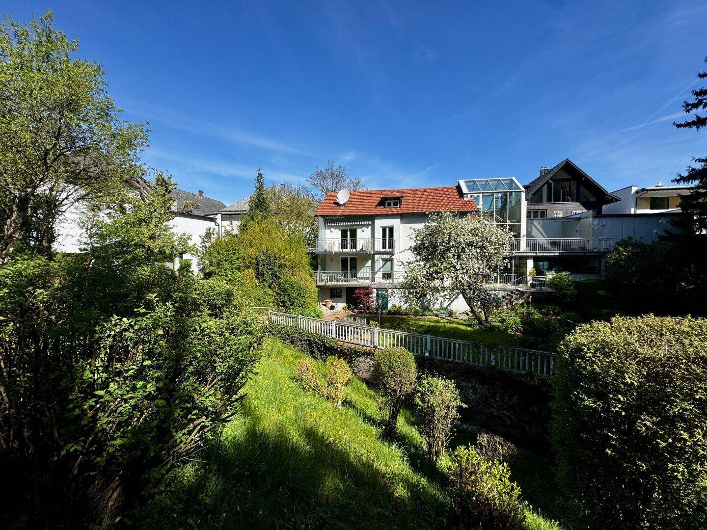 Panoramablick auf das moderne Gebäude mit Glasanbau und terrassiertem Garten unter strahlend blauem Himmel.