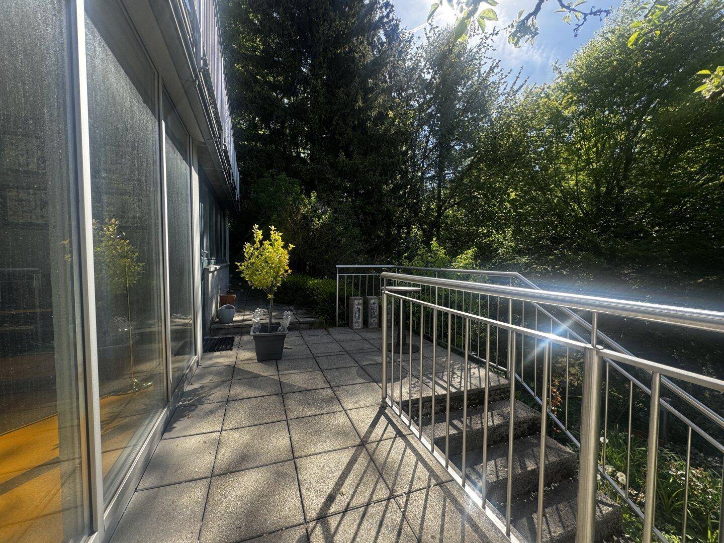 Geräumige geflieste Terrasse mit moderner Glasfassade und Treppenzugang, umgeben von Bäumen.
