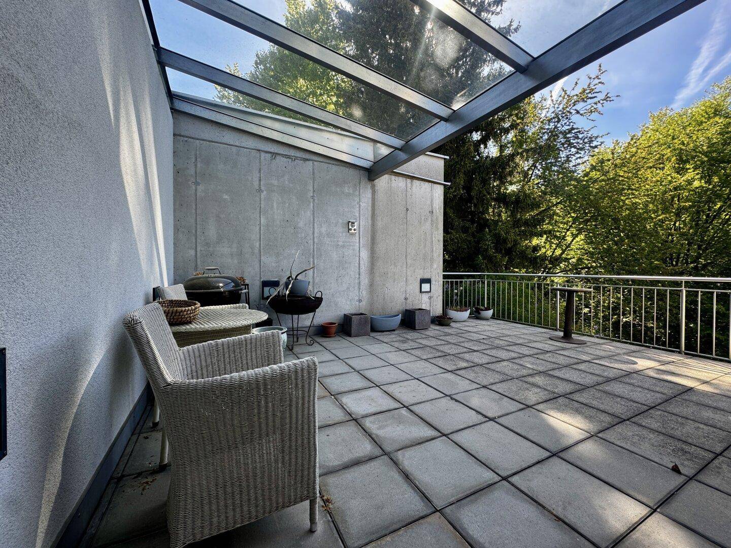 Detailansicht der Terrasse mit Korbsesseln, Grill und Glasüberdachung, umgeben von viel Grün.