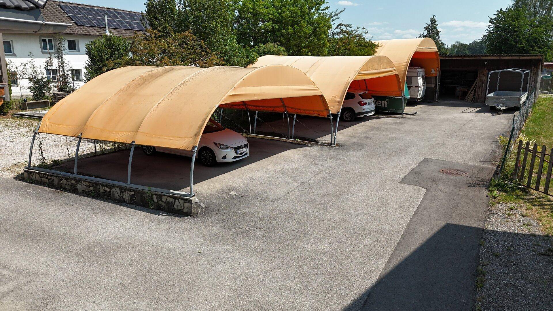 Überdachte Parkplätze mit orangefarbenen Carports auf einem gepflasterten Außenbereich des Grundstücks.