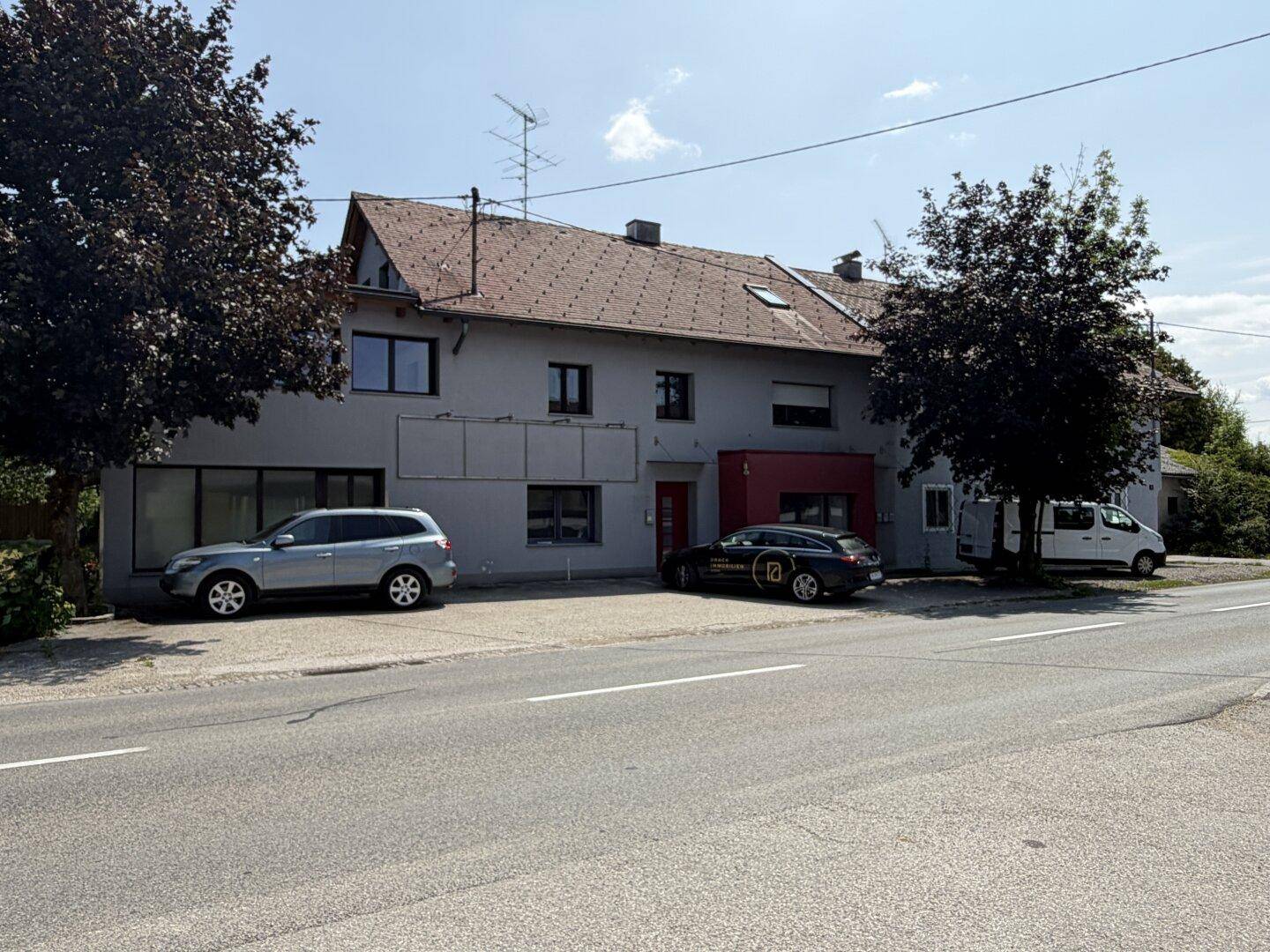 Vorderansicht des Gebäudes mit grauer Fassade, rotem Eingangsbereich und zwei geparkten Autos an der Straße.