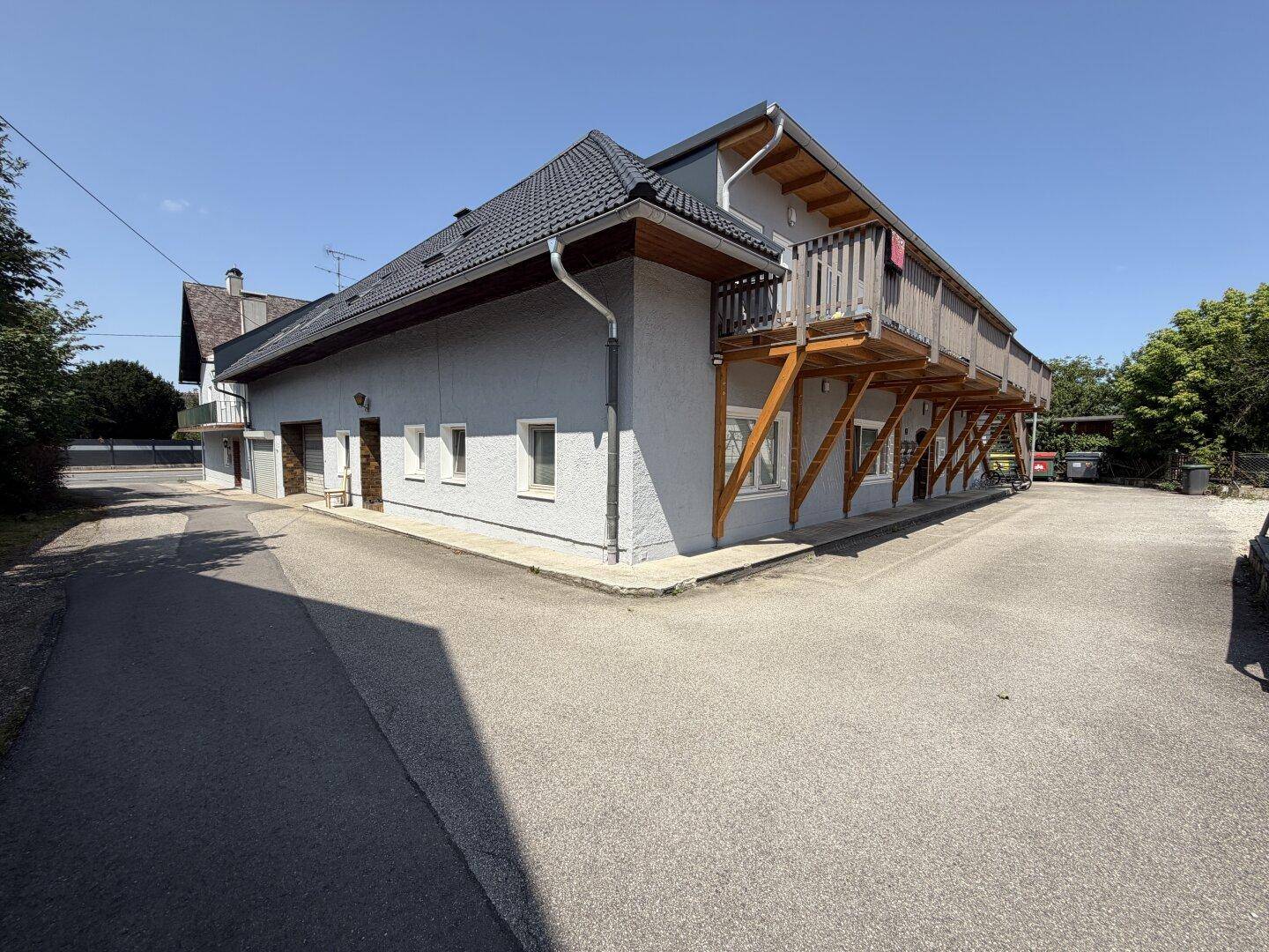 Seitenansicht des Gebäudes mit heller Fassade, dunklem Dach und markantem Holzbalkon über dem Eingangsbereich.
