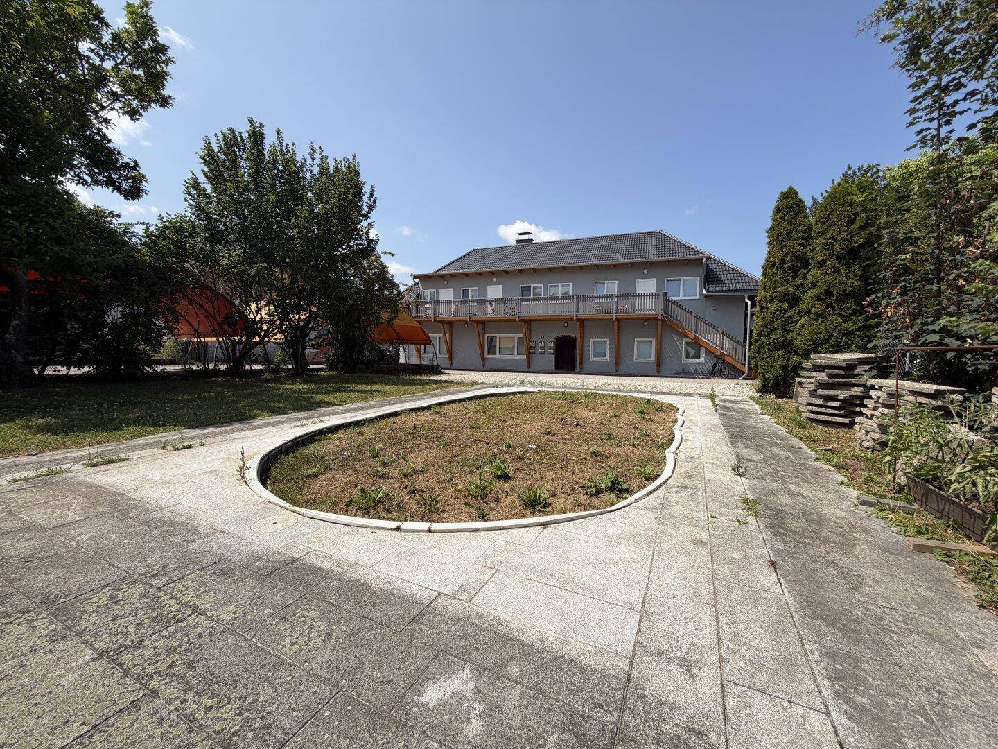 Gepflasterter Vorplatz mit ovaler Rasenfläche und dem Hauptgebäude im Hintergrund, umgeben von Bäumen.