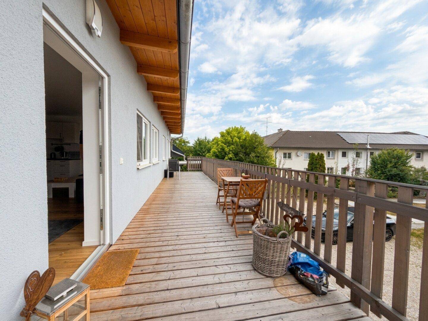 Langer Holzbalkon mit Gartenmöbeln und weitem Blick auf die umliegende Landschaft und Gebäude.