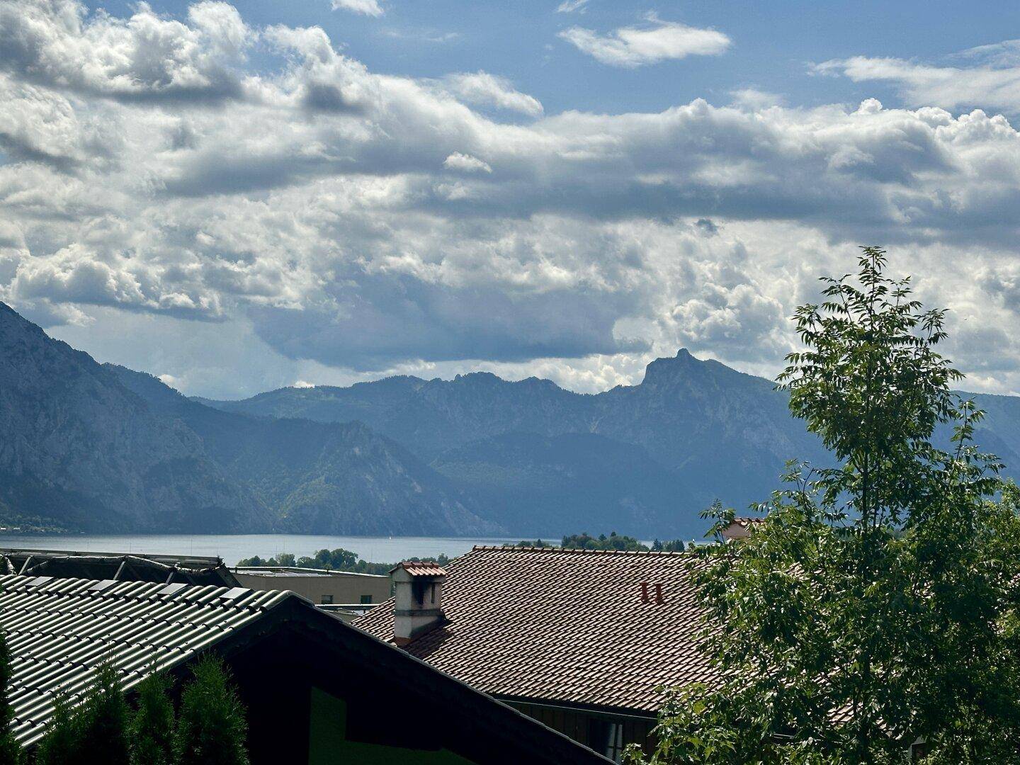 Panoramablick auf einen See und majestätische Berge unter einem bewölkten Himmel, über Dächern und Bäumen.