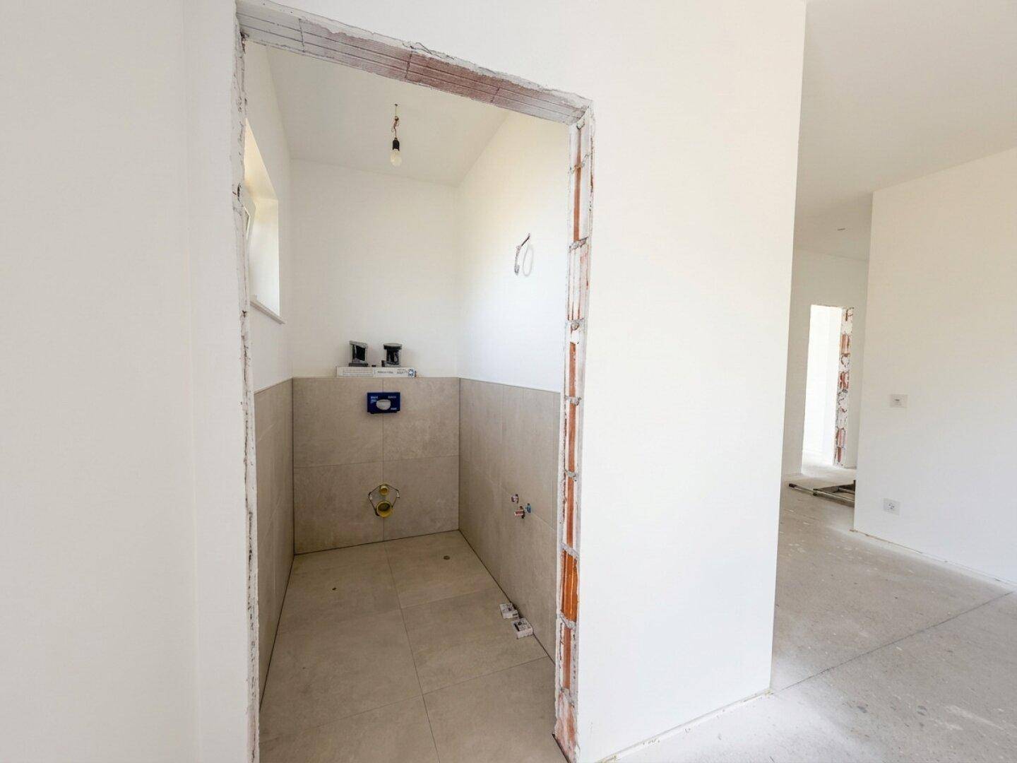 Kleines Gäste-WC im Rohbau mit vorbereiteten Anschlüssen und einem Fenster für Belüftung.