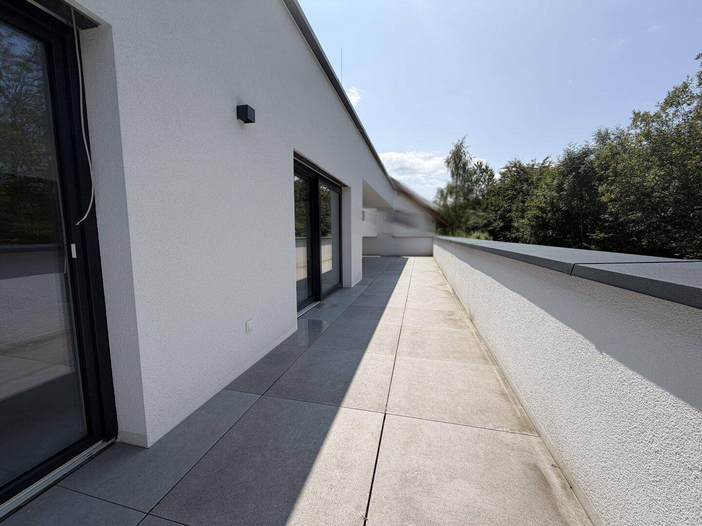 Langer, schmaler Terrassenbereich entlang der modernen Fassade mit Blick ins Grüne.