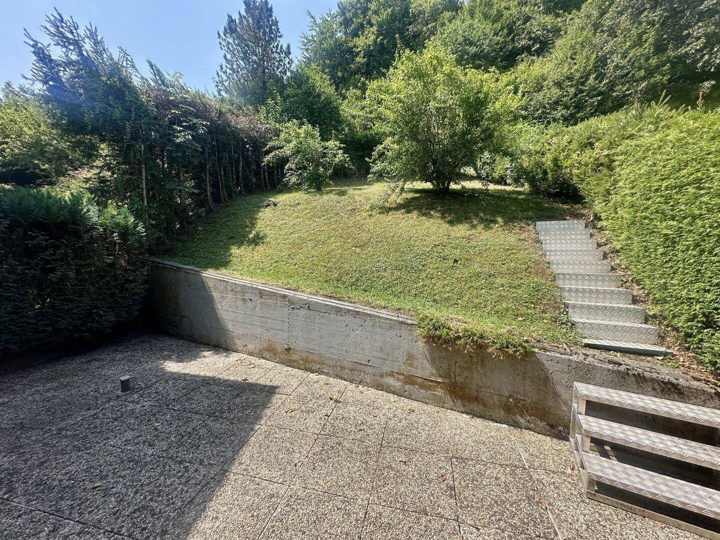 Gepflasterte Terrasse mit Betonmauer und Treppe, die zu einem grünen Gartenhang führt.