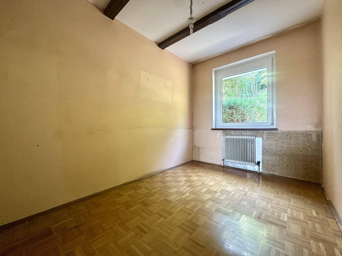 Leeres Zimmer mit Parkettboden, Holzbalken und Fenster mit Blick auf die Natur.